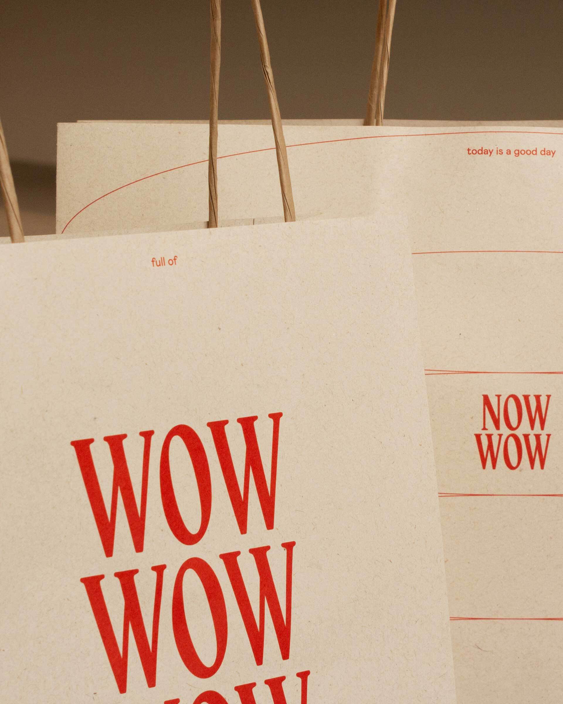 Foto von zwei beige Papiertragetaschen mit rotem Aufdruck. Auf der linken im Vordergrund steht klein "full of", groß "Wow, wow, wow" auf der rechten klein "today is a good day", mittig das Logo und rot linierte Ellipsen.