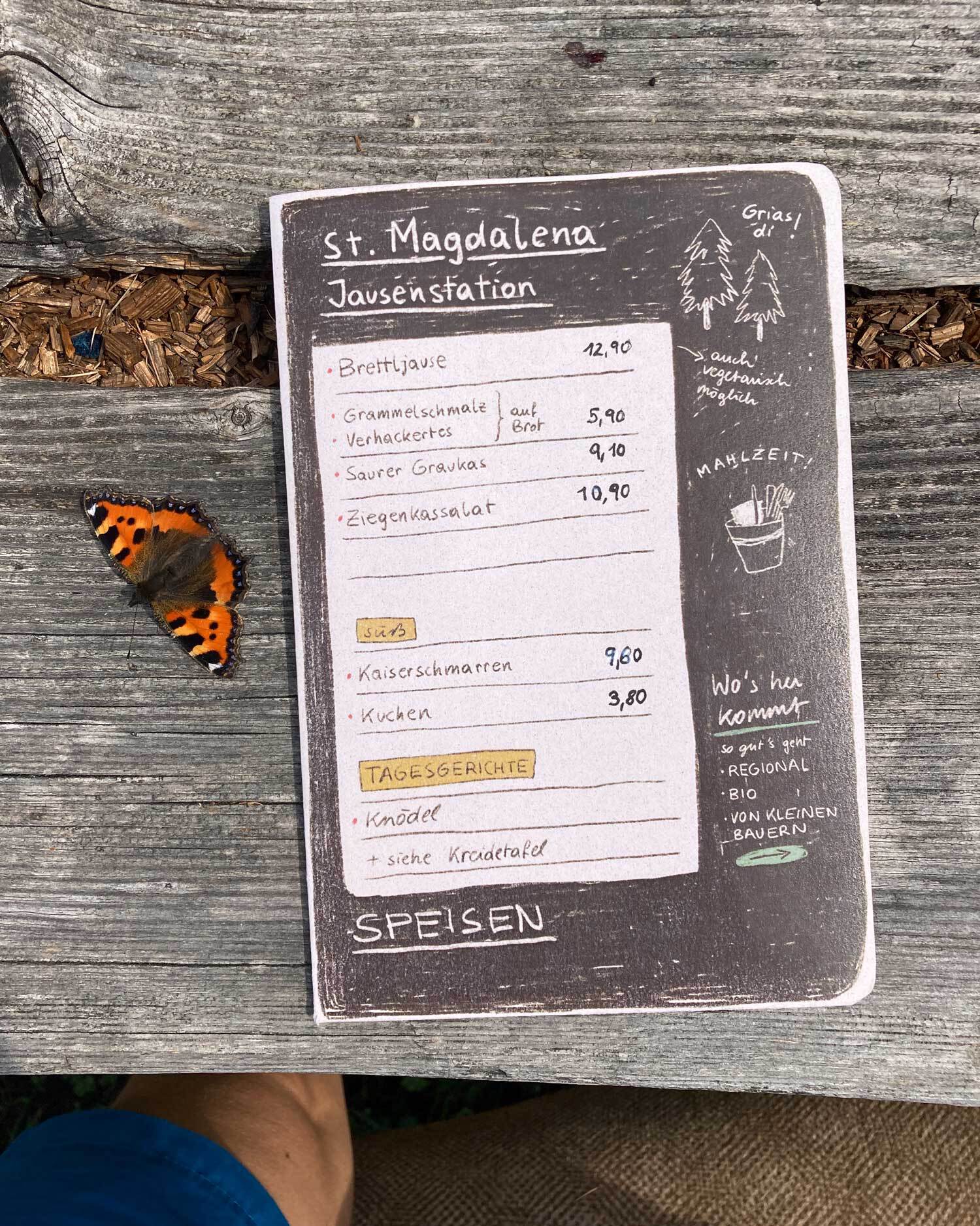 Geschlossene Speisekarte (A5) auf Holztisch. Daneben sitzt ein oranger Schmetterling am Tisch.