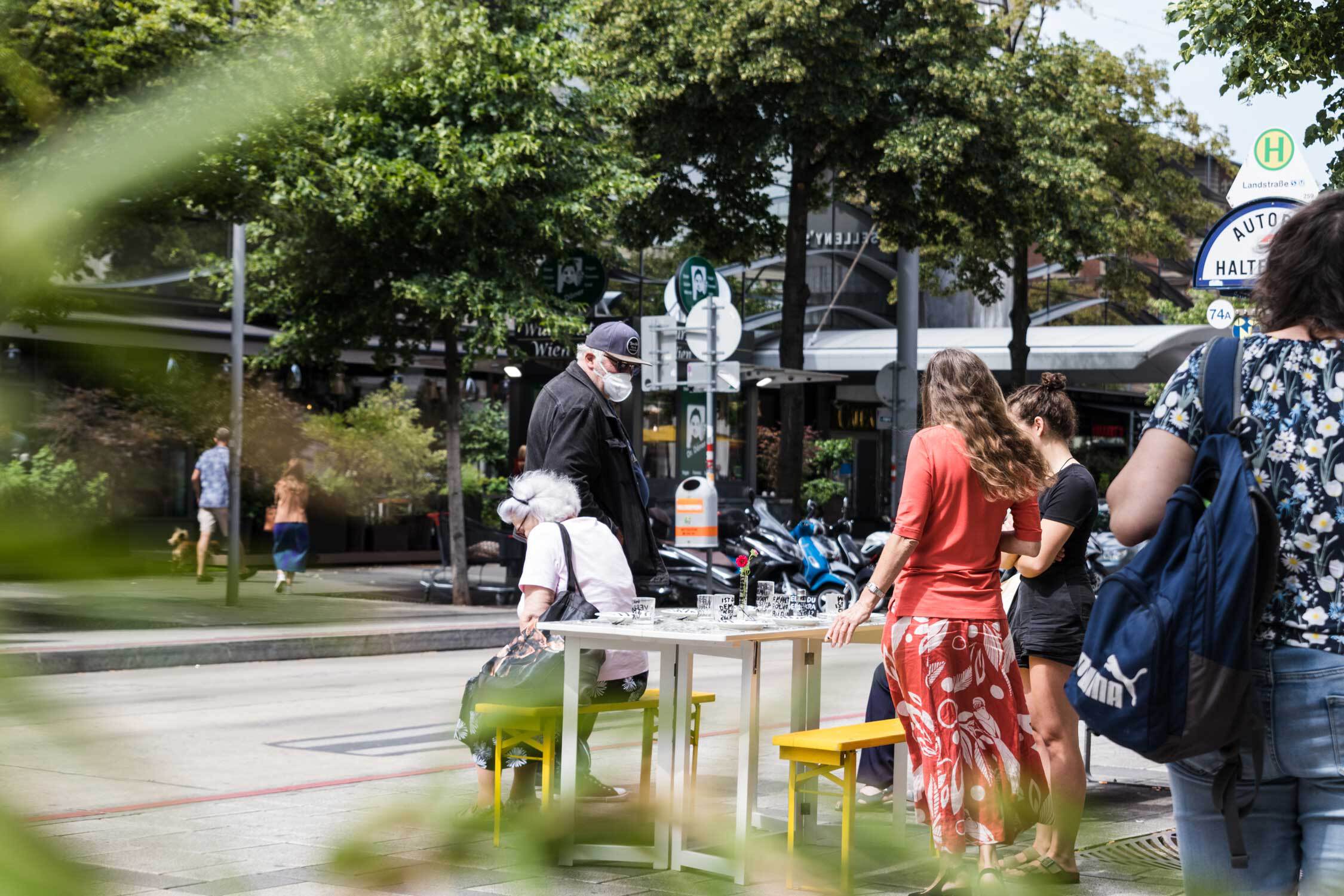 Der Tisch steht aufgeklappt neben einer Bushaltestelle. Ein paar Menschen stehen drum herum und betrachten den Tisch.