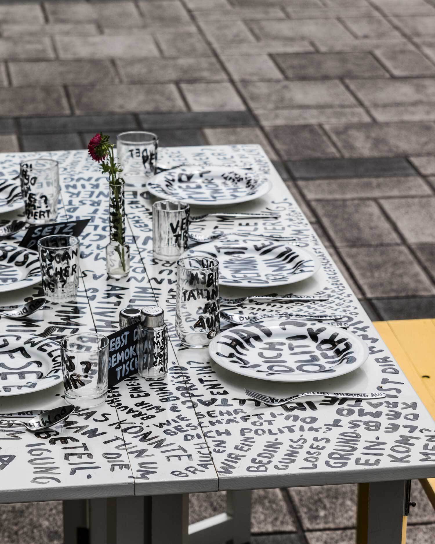 Bild von schräg oben auf den weißen Tisch, mit sechs Gedecken. Alles in Schwarz mit Fragen bepinselt.