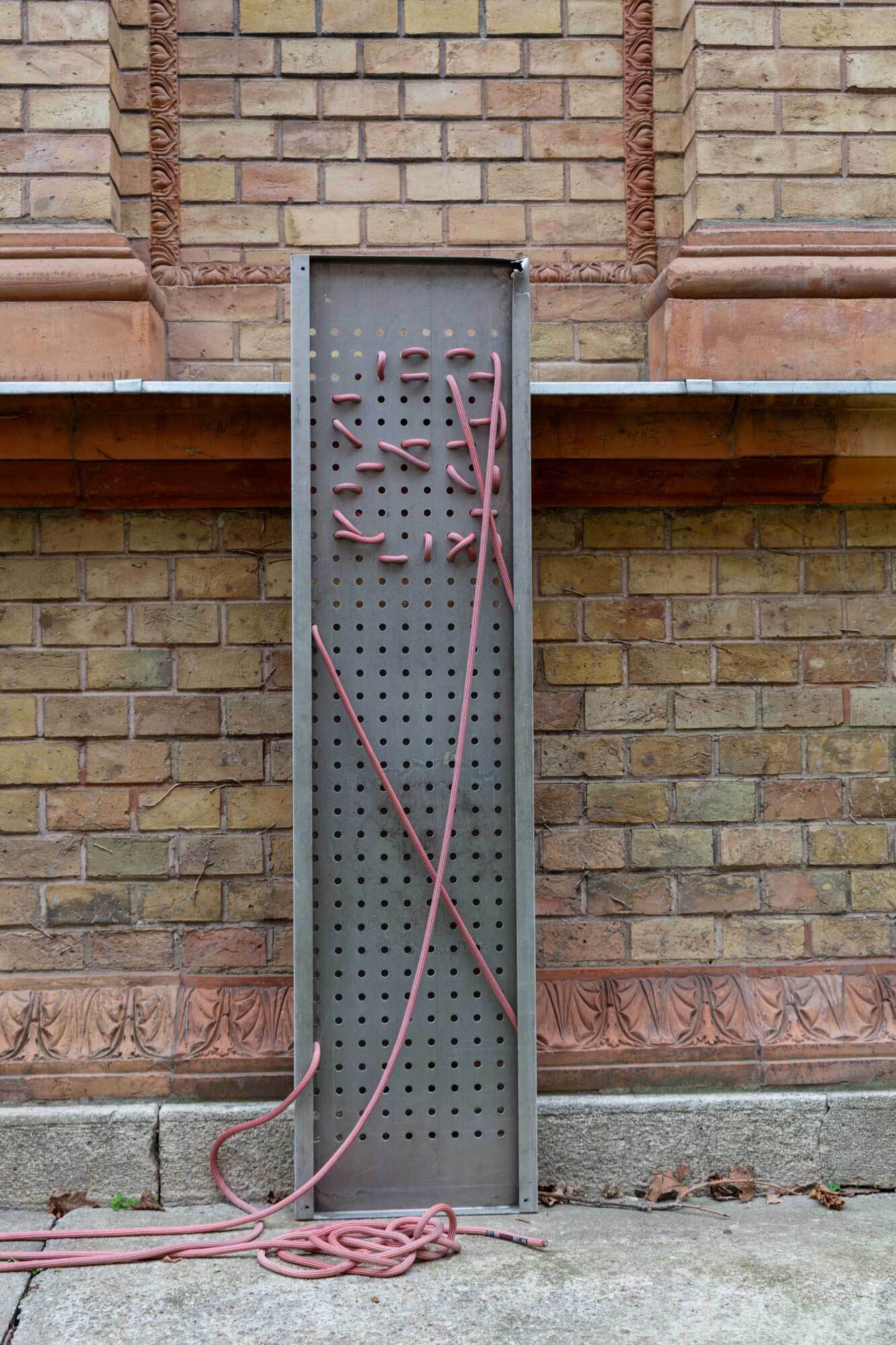 An eine Backsteinwand gelehnes Lochpanel. Durch die Löcher wurde mit einer roten Kletterschnur ein B gewebt.