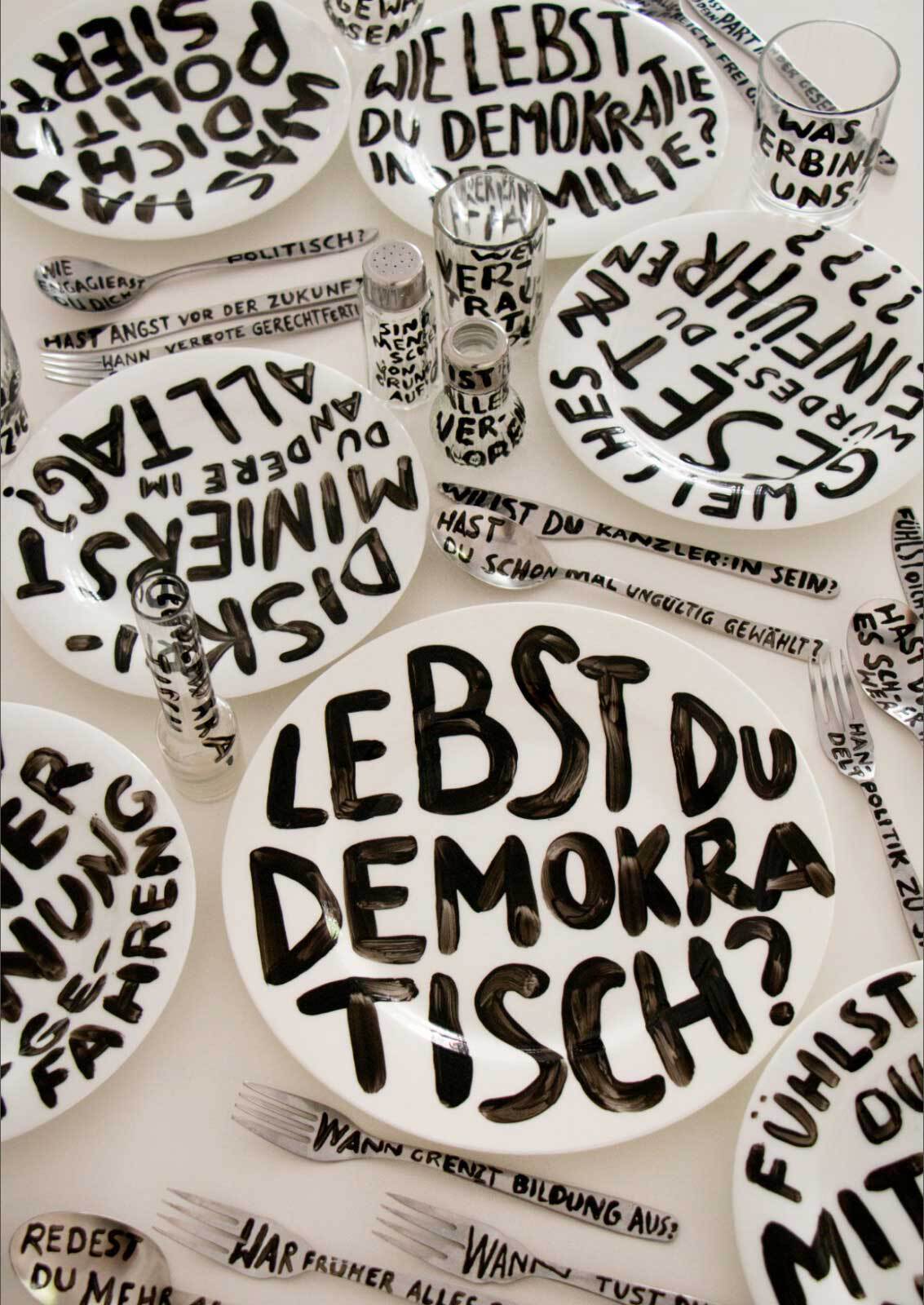 Foto von Tellern, Gläsern und Besteck, auf die mit schwarzer Farbe Fragen gepinselt wurden.